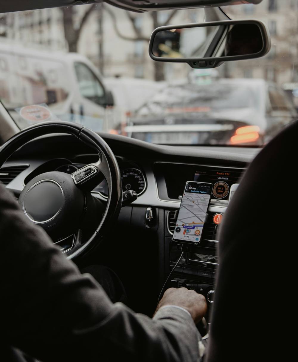 Un chauffeur au volant, vu de l’arrière : sa main sur le levier, un téléphone sur support au tableau de bord, une rue de Paris devant le pare-brise